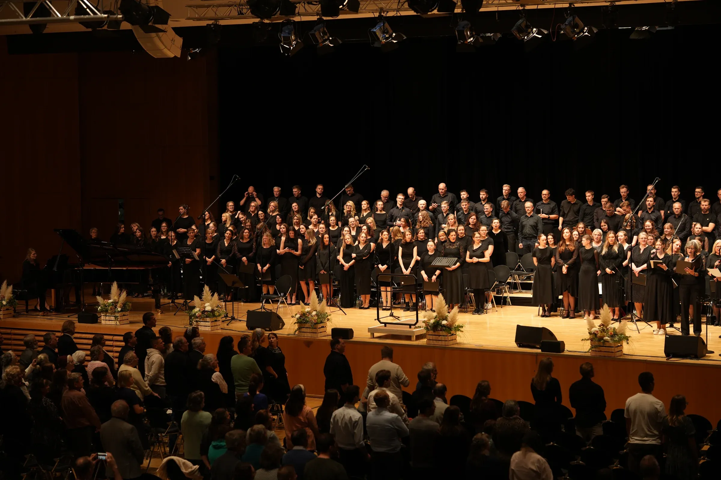 Der große Chor steht zum Abschluss auf der Bühne, davor das Publikum im Saal.
