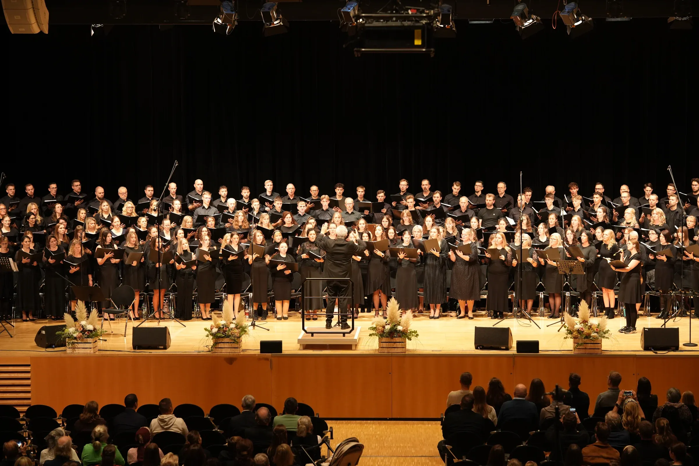 Ein großer Chor in schwarzer Kleidung singt auf der Bühne der Harmonie Heilbronn, vorne der Dirigent, im Saal das Publikum.