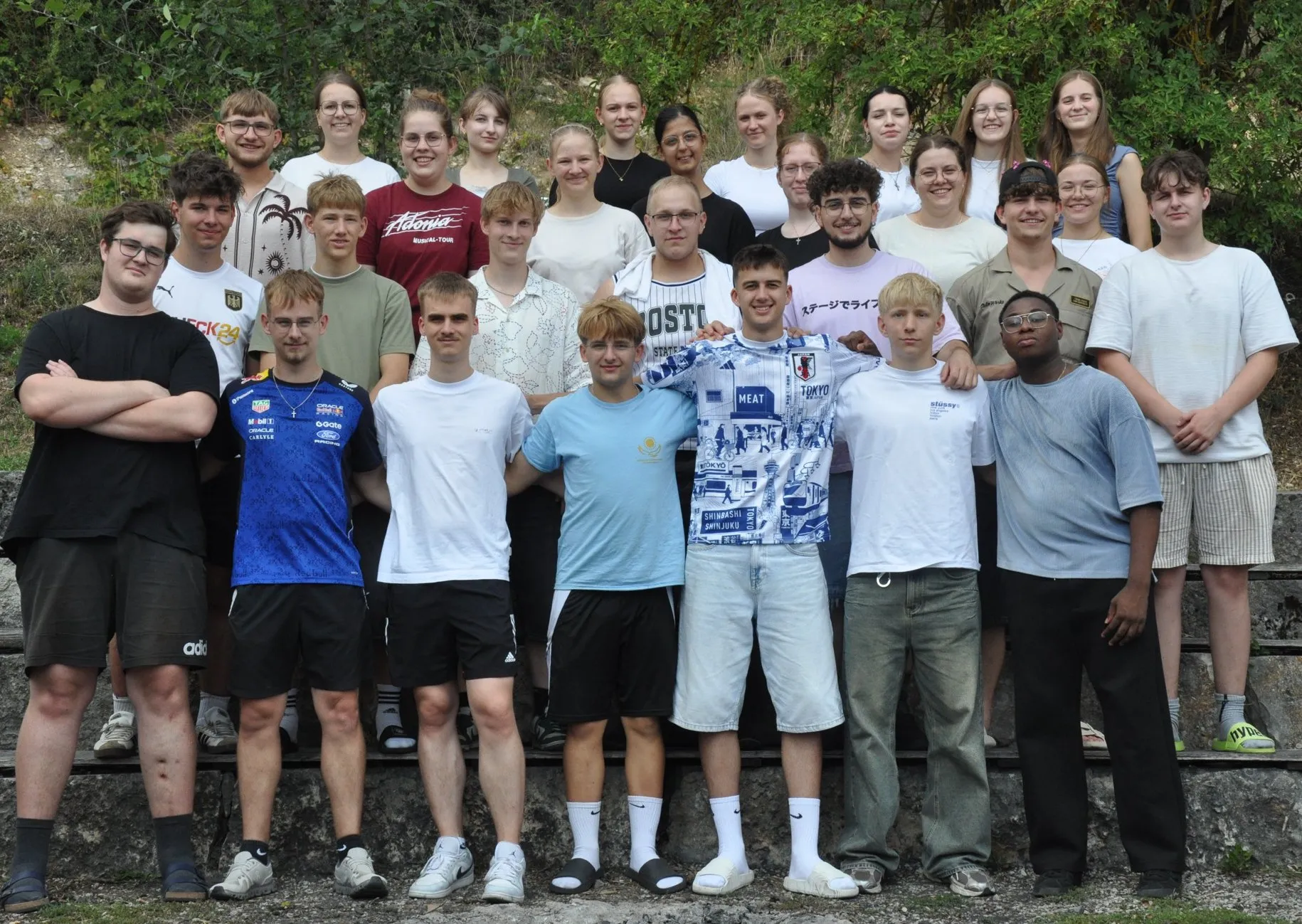 Gruppenfoto von rund 30 jungen Leuten in Sommerkleidung, die lächelnd auf Steinstufen vor grünen Büschen stehen.