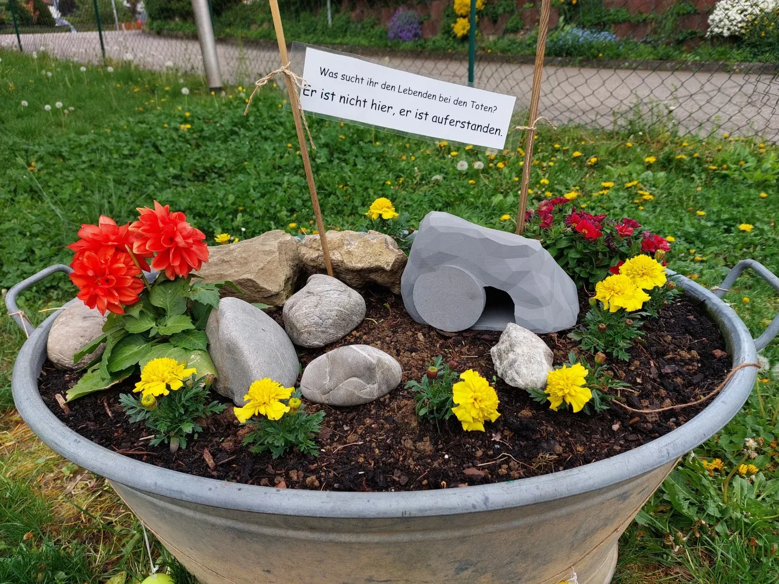 Zinkwanne als Ostergarten mit leerem Grab, Steinen und Blumen, Schild: „Er ist nicht hier, er ist auferstanden.