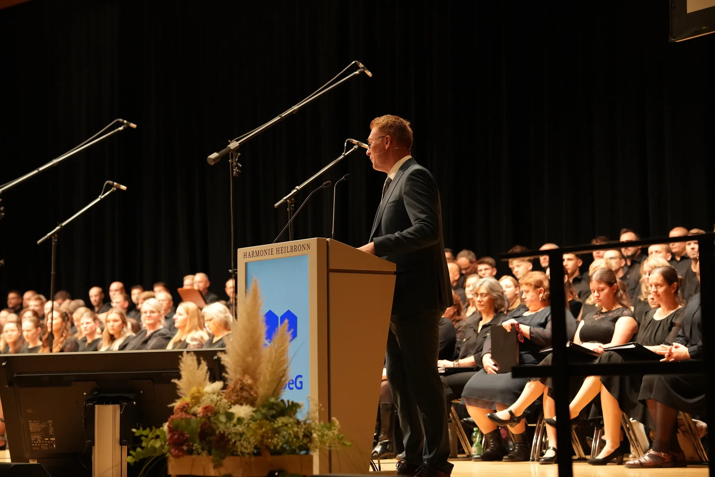 Ein Redner spricht am Pult mit der Aufschrift „Harmonie Heilbronn“, hinter ihm sitzt der Chor.