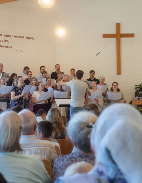 Chor singt vor dem Holzkreuz im Gemeindesaal, die Gemeinde hört zu