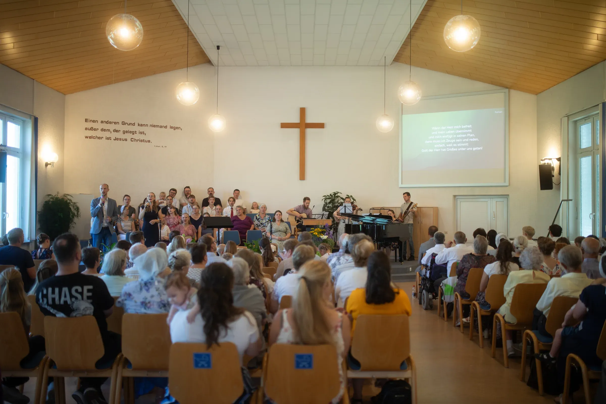 Gottesdienst im Gemeindesaal: Die Gemeinde sitzt in Reihen, vorne das Holzkreuz.