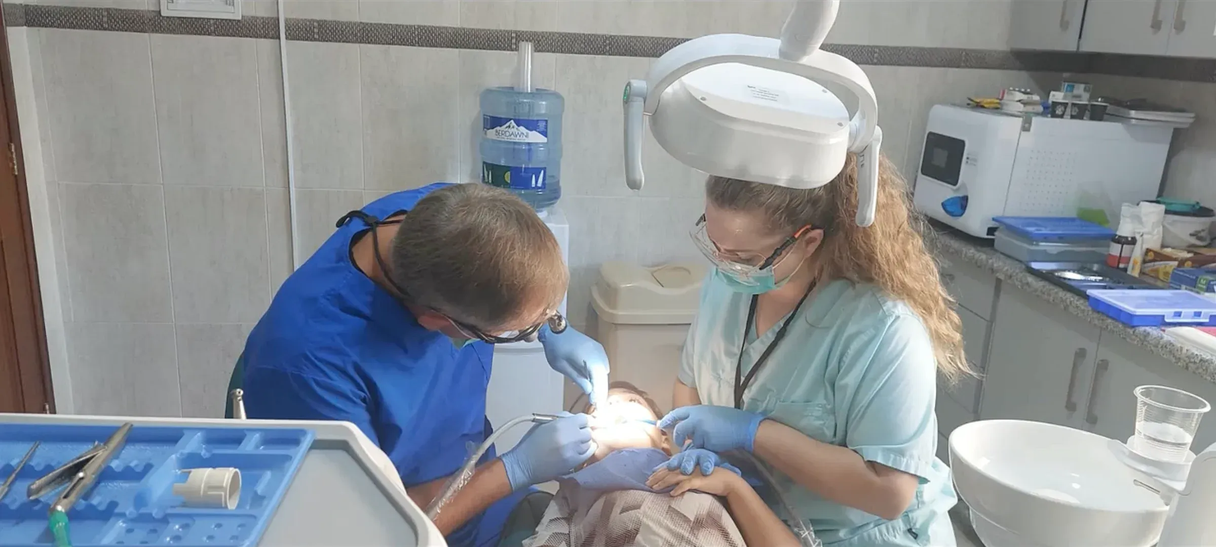Ein Zahnarzt und eine Assistentin mit Mundschutz behandeln eine Patientin unter einer hellen Lampe in einer Zahnarztpraxis.